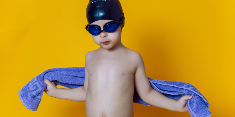 child athlete wearing blue swimming goggles black rubber cap towel stands yellow wall